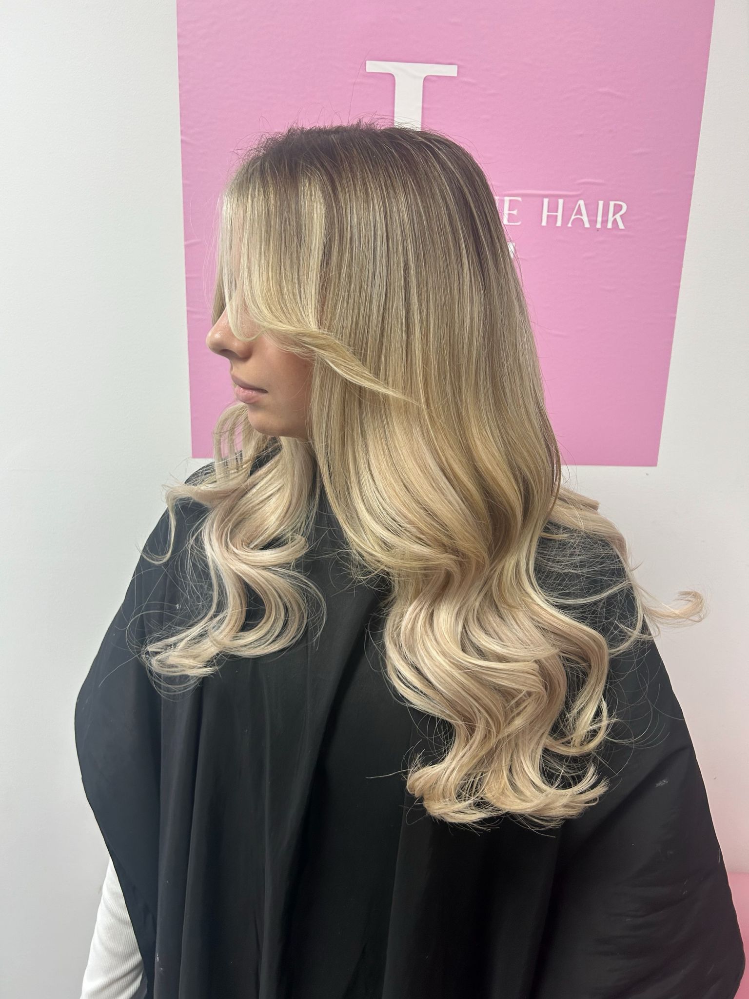 A person with long wavy blonde hair, wearing a black garment, standing in front of a pink wall.